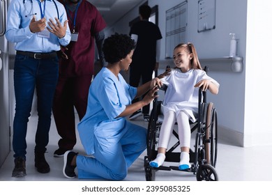 Happy diverse female doctor talking with girl in wheelchair in hospital corridor. Medicine, healthcare, childhood and medical services, unaltered. - Powered by Shutterstock