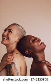 Happy diverse female models standing back to back with their head touching. Two women standing together and smiling over beige background.