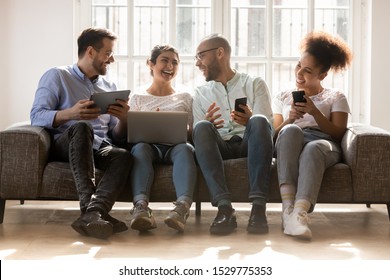 Happy diverse friends using devices, phones, laptop and tablet, having fun together, sharing discussing news, sitting on couch in row, chatting in social network, joking, enjoying meeting - Powered by Shutterstock