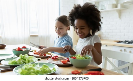 Happy Diverse Little Girls Sisters Preparing Food For Parents, Sitting At Wooden Table In Kitchen Together, Smiling African American And Caucasian Kids Cooking Salad, Chopping Fresh Vegetables