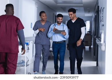 Happy diverse male and female doctors using tablet walking in hospital corridor. Medicine, healthcare, communication and medical services, unaltered. - Powered by Shutterstock