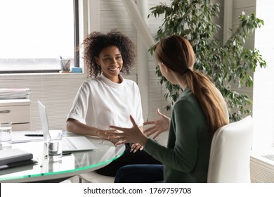 Happy diverse millennial women distracted from computer work chat talk in office during break, smiling young multiracial female colleagues have fun brainstorm discussing ideas or thoughts at workplace