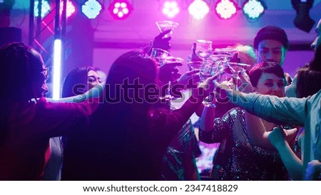 Happy diverse people clinking glasses at disco party, drinking alcohol and saying cheers on dance floor. Men and women partying and having fun at nightclub, raising drinks at event.