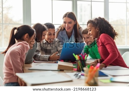 Happy Diverse School Children And Teacher Woman Having Class Sitting At Desk In Classroom At School. Modern Education And Knowledge Concept. Selective Focus