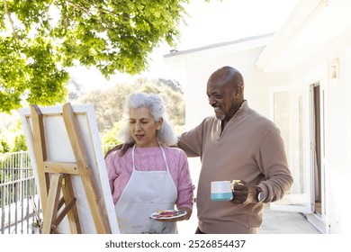 Happy diverse senior couple drinking coffee, embracing and looking at painting on sunny terrace. Lifestyle, retirement, senior lifestyle, nature, creativity, togetherness and domestic life, unaltered. - Powered by Shutterstock