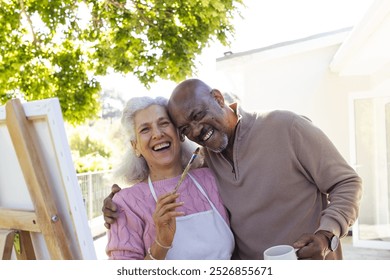 Happy diverse senior couple embracing and looking at painting on sunny terrace. Lifestyle, retirement, senior lifestyle, nature, creativity, togetherness and domestic life, unaltered. - Powered by Shutterstock