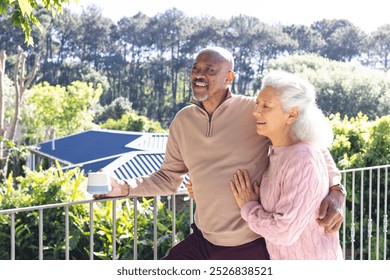 Happy diverse senior couple holding cup of coffee and embracing on sunny terrace. Lifestyle, retirement, senior lifestyle, nature, togetherness and domestic life, unaltered. - Powered by Shutterstock