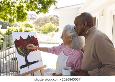 Happy diverse senior woman painting on canvas and senior man embracing her on sunny terrace. Lifestyle, retirement, senior lifestyle, nature, creativity, togetherness and domestic life, unaltered. - Powered by Shutterstock