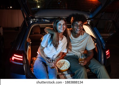 Happy diverse young couple having romantic night. Cheerful guy and his girlfriend watching a movie, sitting together in car trunk in front of a screen in a drive in cinema. Entertainment ideas