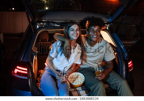 Happy diverse young couple having romantic night.\
Cheerful guy and his girlfriend watching a movie, sitting together\
in car trunk in front of a screen in a drive in cinema.\
Entertainment ideas