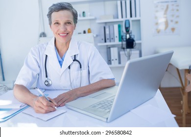 Happy doctor looking at camera and writing on clipboard in medical office - Shutterstock ID 296580842