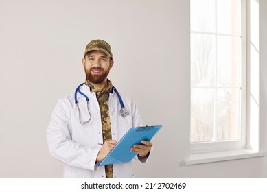 Happy doctor at military hospital. Bearded man wearing camouflage hat and white medical lab coat holding clipboard and pen, looking at camera and smiling. Medicine, healthcare, and army concepts