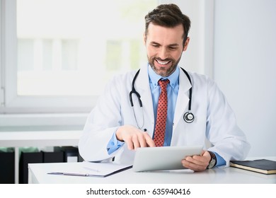 Happy Doctor Sitting At Desk, Using Tablet