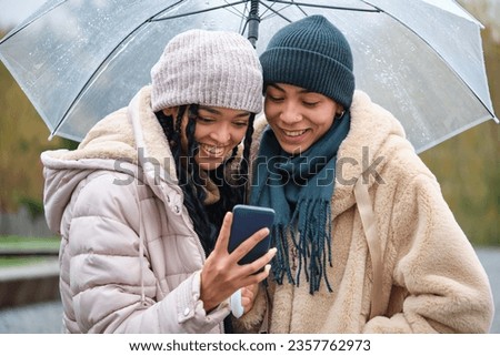 Happy dominican lesbian couple using the smartphone at street in a rainy day of winter.