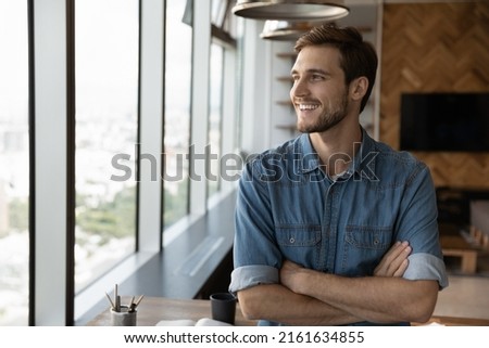 Happy dreaming businessman standing by office or home workplace, looking out of window, thinking of future vision with confidence, smiling at good thoughts, planning investment, enjoying success