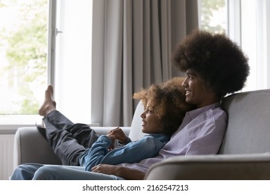 Happy Dreamy African Teen Couple In Love Resting On Couch, Looking Away, Smiling, Laughing, Talking, Dreaming. Young Teenage Black Boyfriend With Long Curly Hair Hugging Girlfriend, Enjoying Romance