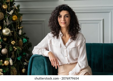 Happy, dreamy, curly-haired black-haired woman sits near the Christmas tree, gazing into the distance, making a wish. Smiling young woman enjoying Christmas at home sitting on the couch