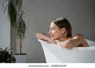 Happy, dreamy girl with haircut resting in bathtub, enjoying pastime in spa. Close up of relaxed caucasian woman leaning, putting crossed arms on bath edge and smiling. Wellness, spa concept.