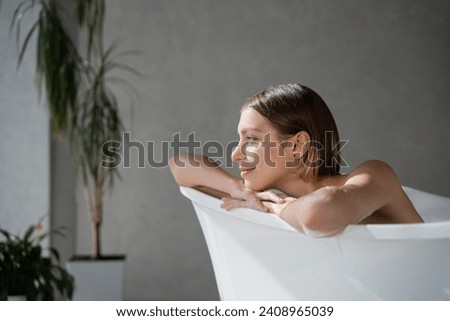 Happy, dreamy girl with haircut resting in bathtub, enjoying pastime in spa. Close up of relaxed caucasian woman leaning, putting crossed arms on bath edge and smiling. Wellness, spa concept.