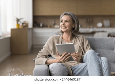 Happy dreamy middle-aged woman holds digital tablet, sitting on cozy sofa, looking away, chatting in social media, enjoy online communication, thinking, planning, using on-line application for hobby - Powered by Shutterstock