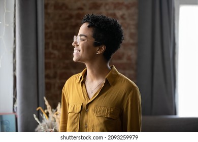 Happy Dreamy Millennial African American Woman In Eyeglasses Looking In Distance, Imagining Good Future, Remembering Positive Life Moments, Feeling Inspired Posing In Modern Loft Style Home Or Office.