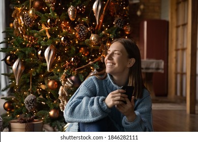 Happy dreamy millennial woman sit near Christmas tree use cellphone look in distance making wish. Smiling young female enjoy New Year winter holiday at home, dreaming or visualizing of joyful future. - Powered by Shutterstock