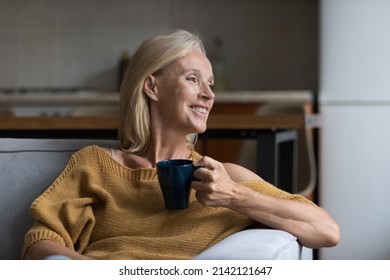 Happy dreamy senior blonde lady enjoying good morning, drinking hot coffee at home, resting on comfortable couch in cozy living room, holding ceramic mug, cup of tea, smiling, looking away - Powered by Shutterstock