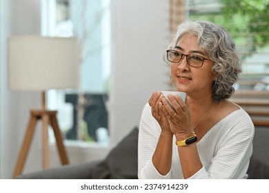 Happy dreamy Senior middle-aged Asian woman with eyeglasses sitting on a comfortable sofa in the living room with a cup of coffee, looking away, Peaceful, and enjoying activity alone at home..