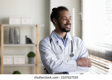 Happy Dreamy Young African American Male Doctor Physician Therapist In White Coat Standing With Folded Arms In Clinic, Looking In Distance, Thinking Of Career Opportunities Or Medical Challenges.