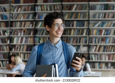 Happy dreamy young Jewish male student in eyewear holding books and cellphone in hands, looking in distance in library thinking of getting message with pleasant news or email with good test results.