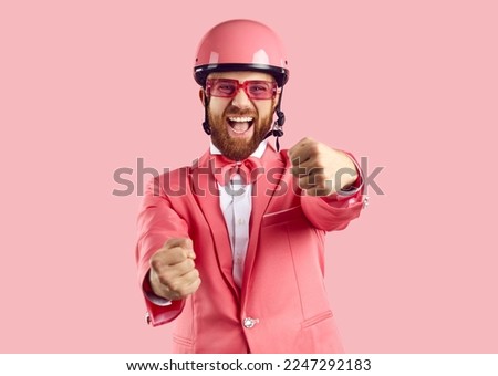 Happy driver holding invisible steering wheel. Studio shot of funny excited man with ginger beard, wearing pink helmet, funky suit, bow tie and glasses having fun and pretending to drive modern car