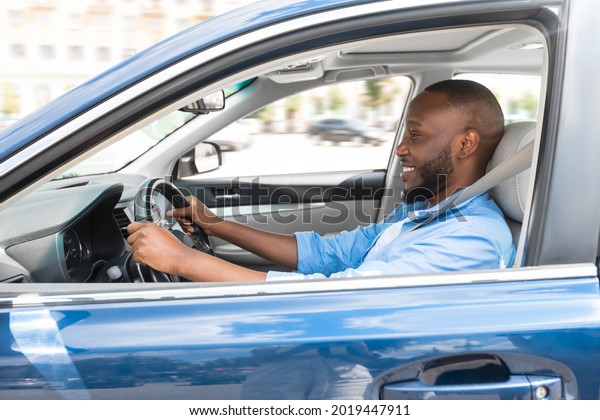 Happy\
Driver. Side view profile portrait of cheerful positive African\
American man sitting in a car on driver\'s seat. Excited black guy\
riding in the city, holding hands on steering\
wheel