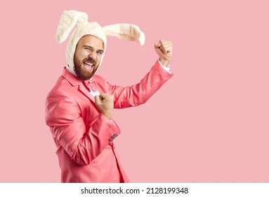 Happy Easter Bunny celebrating success. Funny cheerful confident ginger man wearing white fluffy rabbit ears and funky pink suit standing on pink background, fist pumping and shouting Yes, Yay, Hooray