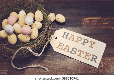 Happy Easter candy easter eggs in birds nest on dark vintage recycled wood background with Happy Easter gift tag sign message greeting, with applied retro style filters.  