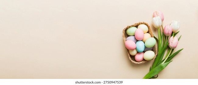 Happy Easter composition. Easter eggs in basket on colored table with yellow Tulips. Natural dyed colorful eggs background top view with copy space.