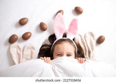 Happy Easter. cute beautiful girl with blue eyes and bunny ears peeking out from under the blanket. Chocolate eggs are scattered on the bed. Egg hunt. Lifestyle High quality photo