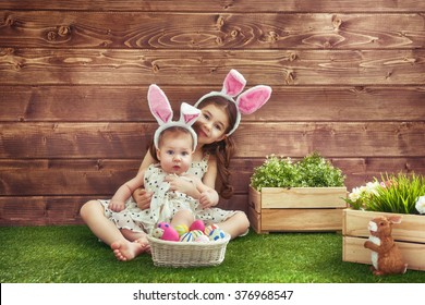 Happy Easter! Cute Little Children Girls Wearing Bunny Ears On Easter Day. Sisters Hunting For Easter Eggs On The Lawn Near The House. Girls Having Fun, Playing And Hugging.