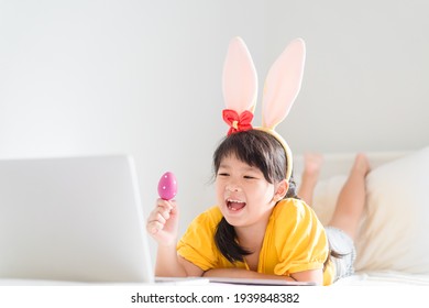 Happy Easter Day.Asian Child Girl In Rabbit Ears Holding Eggs.Online Worship In Easter Holidays With Laptop.Kid Child Online With Bunny Hat, Easter Egg Hunt Online At Home.Lockdown, Covid19, Medical.