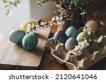 Happy Easter! Easter eggs on rustic table with cherry blossoms. Natural dyed colorful eggs in paper tray on wooden board and spring flowers in rustic room. Moody atmospheric image