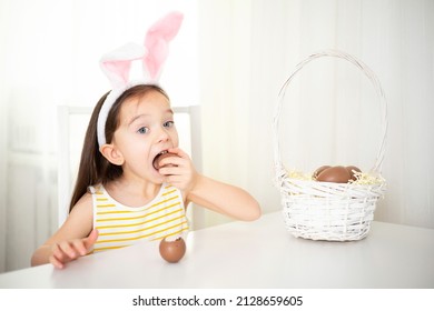 Happy Easter. A funny cute girl with bunny ears eats a chocolate egg at the table with pleasure. Lifestyle. Light colors. emotions High quality photo