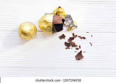 Happy Easter. Stylish Easter Egg In Golden Foil And Broken Chocolate Egg With Chocolate Pieces On White Wooden Background, Space For Text. Modern Gold Easter Eggs. Holiday Gift