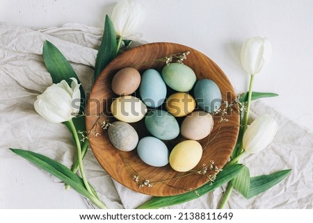 Happy Easter! Stylish Easter eggs in wooden plate, tulips and linen napkin on rustic white table, flat lay. Natural dyed colorful eggs and spring flowers rustic composition.