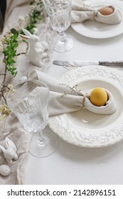 Happy Easter! Stylish elegant Easter brunch table setting. Easter egg in bunny napkin on plate with cutlery, bunny, flowers on rustic white table. Modern eco table arrangement. Top view