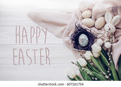 happy easter text greeting card sign on  stylish easter eggs in nest with bunny on beige fabric and tulips and willow buds on rustic white wooden background flat lay. space for text, top view