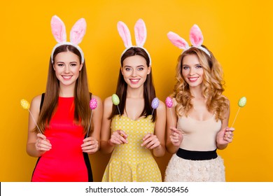 Happy Easter! Three Pretty, Trendy Girls Wearing Bunny Ears On Heads In Dresses Holding Colorful Easter Eggs, Standing Over Yellow Background