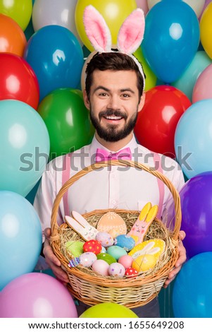 Happy easter. Vertical photo of funny guy colorful decor party mood hold painted eggs ginger bread basket wear pink shirt bow tie suspenders bunny ears on balloons background