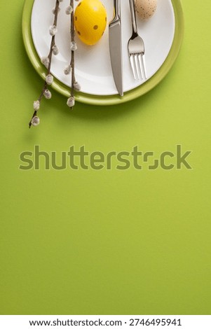 Happy Easter vertical table setting featuring a painted yellow egg, speckled egg, pussywillow branches and stainless cutlery on layered plates over a green background ideal for spring holiday visuals