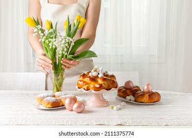 Happy Easter! Woman preparing table with Easter pastry, eggs, candies and spring flowers for holiday. Light background. Copy space.