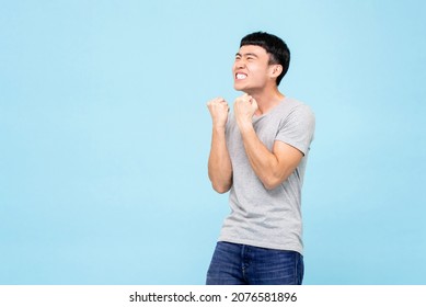 Happy ecstatic young Asian man raising his fists doing yes gesture celebrating success in isolated light blue studio background
