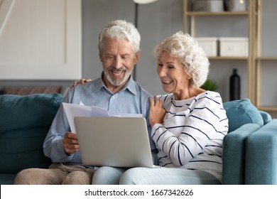 Happy Elderly 60s Couple Sit Rest On Couch At Home Pay Household Expenses Online On Computer, Smiling Mature 50s Husband And Wife Clients Hold Documents Make Payment On Internet Banking Service